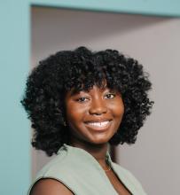 A Black woman with shoulder length curly hair wearing a grey-green sleeveless dress smiles at the camera. 