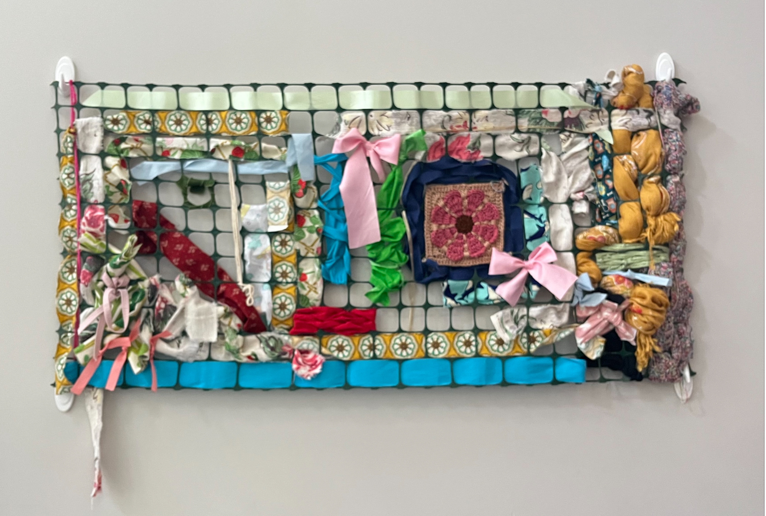 A photo of colorful fabrics, yarn, and crochet pieces woven together on a section of garden fencing, mounted to the wall with hooks